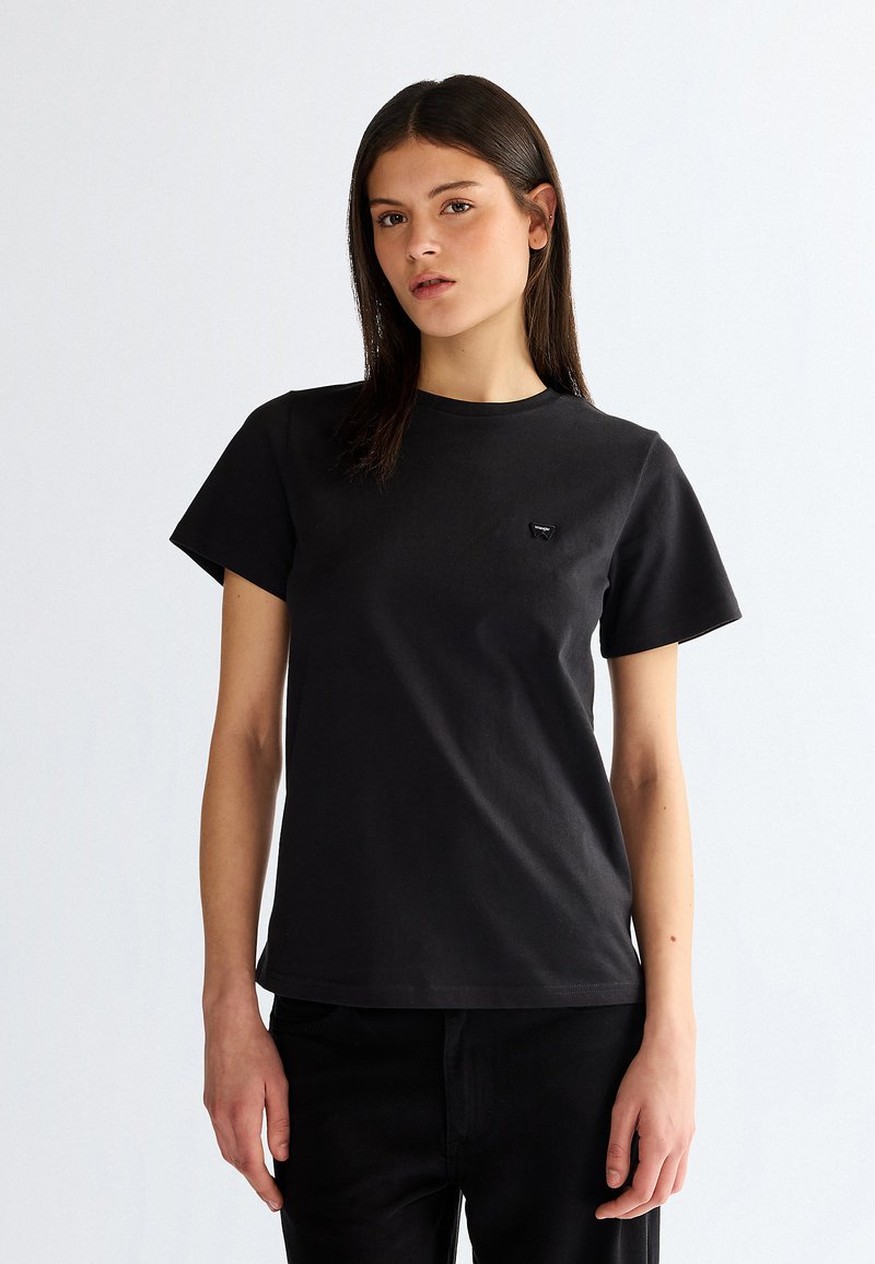 Młoda kobieta z prostymi ciemnymi włosami nosi prostą czarną koszulkę z krótkim rękawem oraz czarne spodnie, stoi na białym tle.