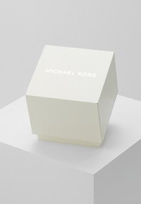 Scatola bianca con texture e cima inclinata, con la scritta "MICHAEL KORS" stampata in bianco sul davanti. Design semplice e geometrico.