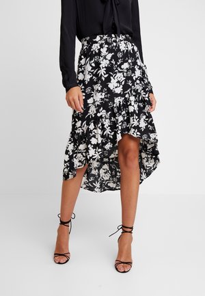 Mujer con falda asimétrica con volantes florales en blanco y negro, blusa negra y sandalias negras de tacón con tiras sobre un fondo liso.