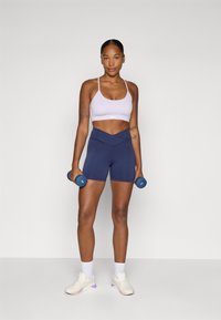 Îmbrăcăminte de antrenament cu un sutien sport violet deschis și șorturi înalți, de culoare bleu marin. Modelul ține gantere albastre deschise și poartă adidași albi.