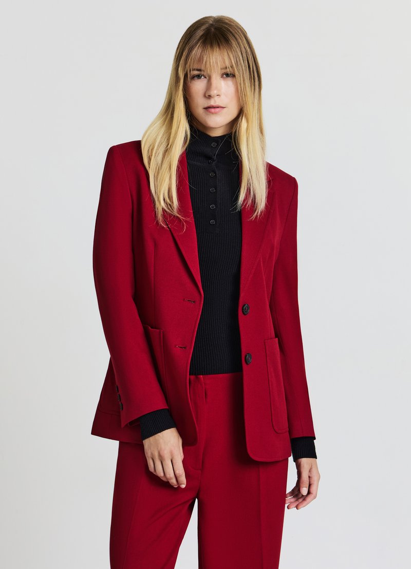 Blazer rosso con vestibilità sartoriale, rever a incavo e tasche anteriori, indossato sopra un dolcevita nero a coste. Tessuto liscio, design minimalista.