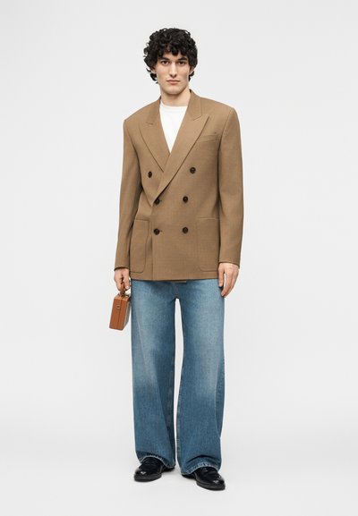 Άνδρας με σγουρά μαλλιά που φοράει καφέ διπλόμπουκο σακάκι, λευκό πουκάμισο, παντελόνι με φαρδιές μπατζάκια, μαύρα παπούτσια, κρατώντας μια μικρή καφέ τσάντα χειρός.