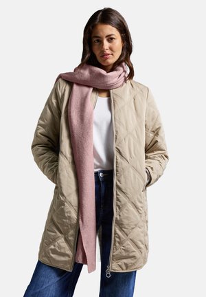 Giacca beige trapuntata con zip, caratterizzata da motivi a diamante. Indossata sopra una camicia bianca e abbinata a una sciarpa rosa in maglia. Jeans blu scuro.