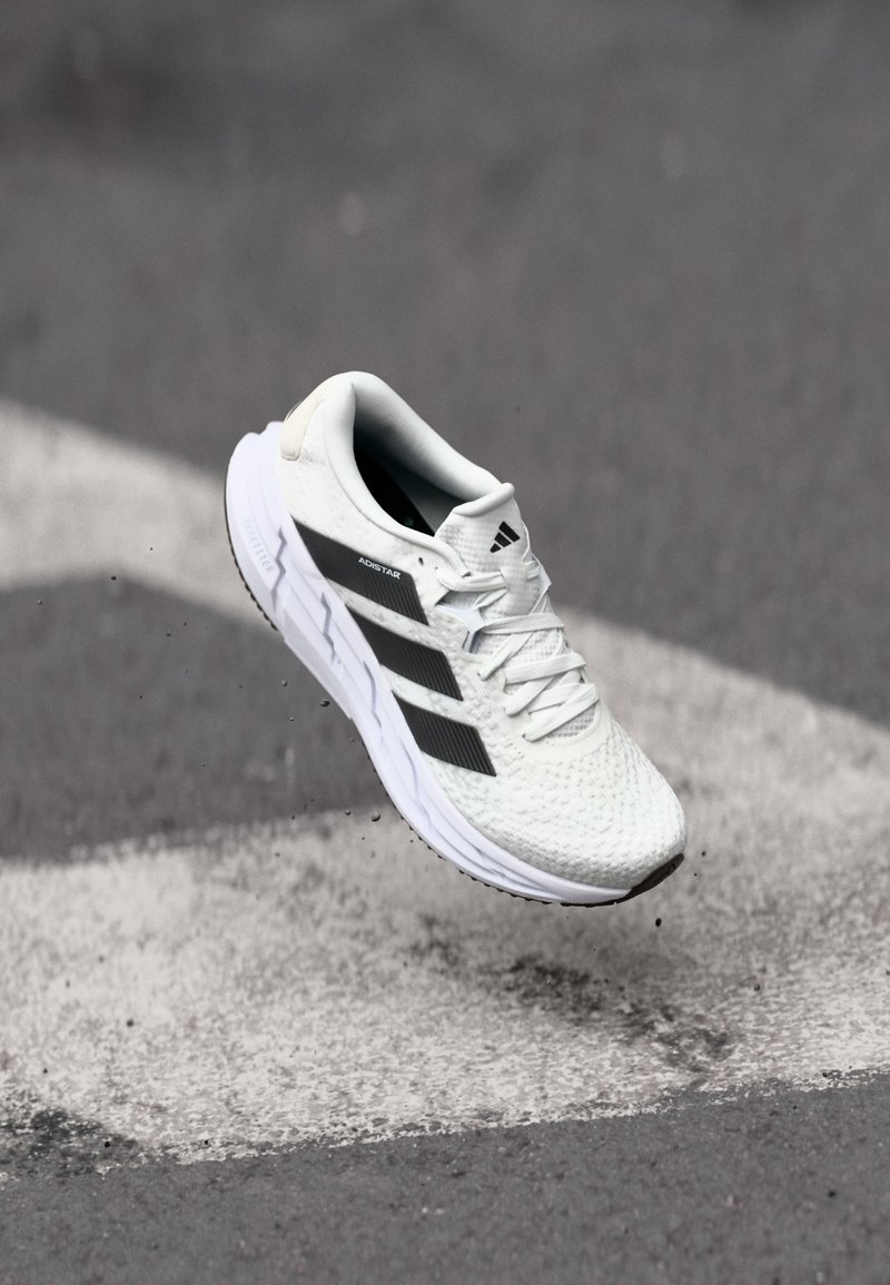 Baskets de sport blanches avec un dessus en mesh texturé, des rayures noires, un laçage et une semelle rembourrée, lévitant au-dessus d'une surface de route marquée.