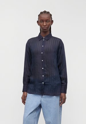 Femme aux cheveux tressés portant une chemise boutonnée bleu marine transparente et un jean bleu clair, debout devant un fond gris clair uni.