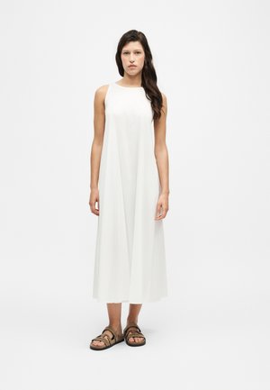 Femme aux longs cheveux foncés portant une robe midi blanche sans manches et des sandales marron à double bride, debout devant un fond blanc uni.