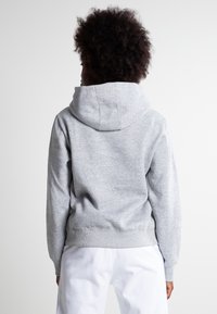 Sudadera gris con una gran capucha, puños y dobladillo acanalados, textura suave, combinada con pantalones blancos, vista desde la parte de atrás.