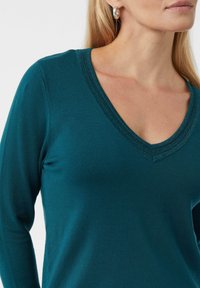 Kobieta w turkusowym swetrze z dekoltem w serek i srebrnych kolczykach w kształcie kółek, odsłaniająca szyję, podbródek i część twarzy na tle białym.