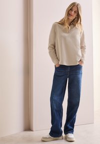 Top de manga larga beige con cuello, combinado con jeans de pierna ancha de color azul oscuro. La modelo lleva zapatillas blancas. Diseño sencillo y de corte relajado.