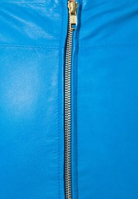 Vêtement en cuir bleu vif avec une fermeture éclair dorée proéminente qui court verticalement, présentant une finition texturée et des coutures lisses.