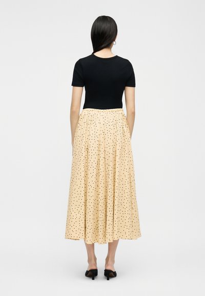 Γυναίκα με μακριά σκούρα μαλλιά, που φοράει μαύρο κοντομάνικο τοπ, μπεζ μίντι φούστα με μαύρες πουά βούλες και μαύρα παπούτσια με τακούνι, που στέκεται γυρισμένη με την πλάτη.