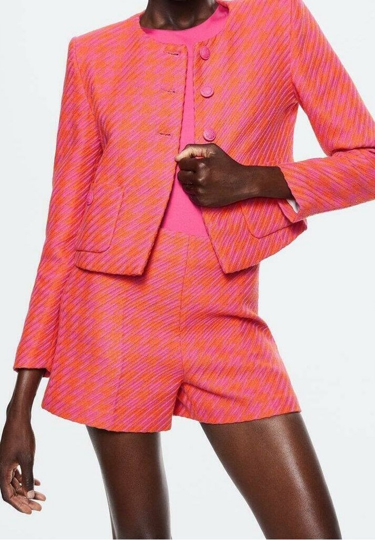 Femme portant un blazer à motif pied-de-poule rose vif et orange, assorti à un short taille haute, avec un haut uni rose en dessous.