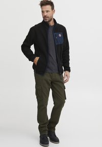 Chaqueta de forro polar negra con detalles azul y cremallera frontal, combinada con pantalones cargo verdes y botas negras. Diseño sencillo y práctico.