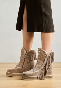 Bottes beiges à talon compensé avec des franges et des strass décoratifs, dotées d'une fermeture éclair sur le côté et d'un dessus en daim texturé avec une semelle épaisse et texturée.