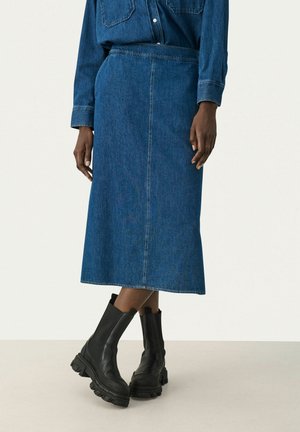 Personne portant une jupe midi en jean bleu, une chemise en jean assortie et des bottines noires en cuir épais sur un fond neutre.
