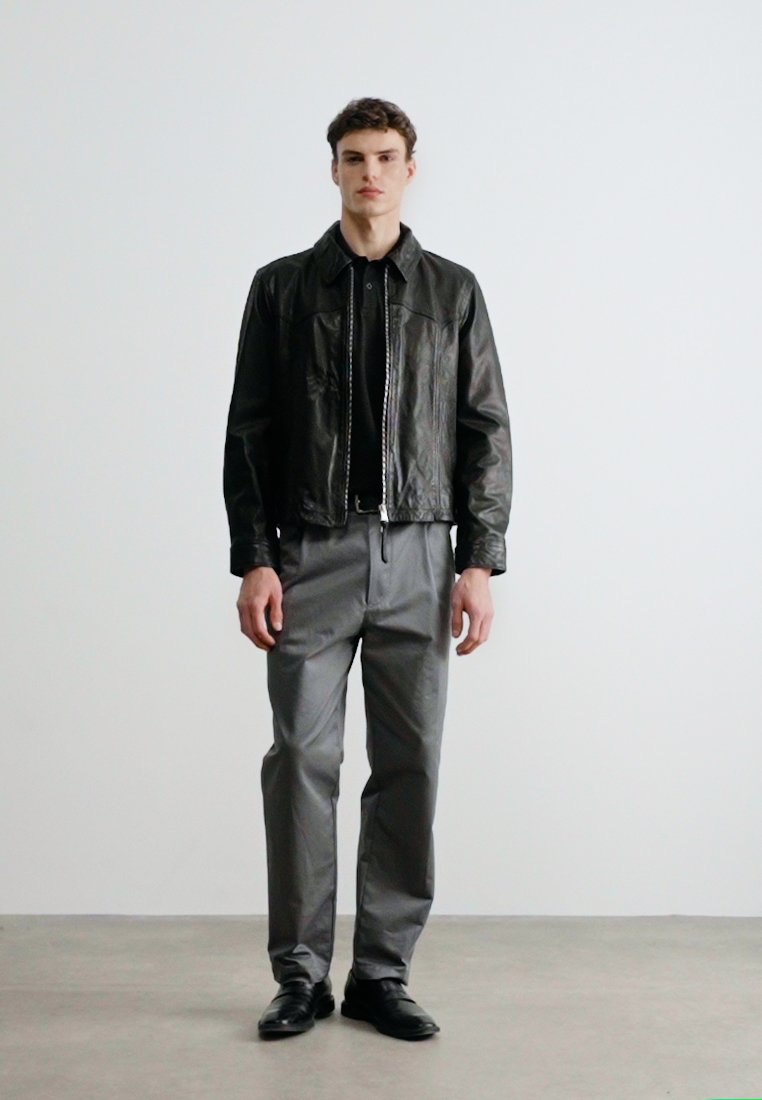 Jeune homme debout contre un mur uni, portant une veste en cuir noire, une chemise noire, un pantalon gris et des chaussures noires, faisant face à l'avant avec une expression neutre.