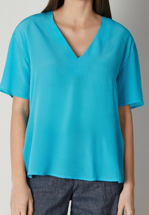 Donna che indossa una blusa turchese a maniche corte e scollo a V, abbinata a pantaloni grigio scuro, in piedi contro uno sfondo semplice.