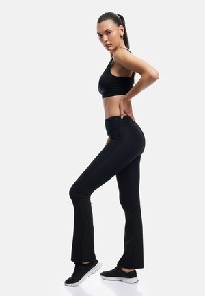 Kobieta w czarnym sportowym biustonoszu, legginsach z wysokim stanem i butach sportowych, stojąca bokiem z jedną ręką na dolnej części pleców.