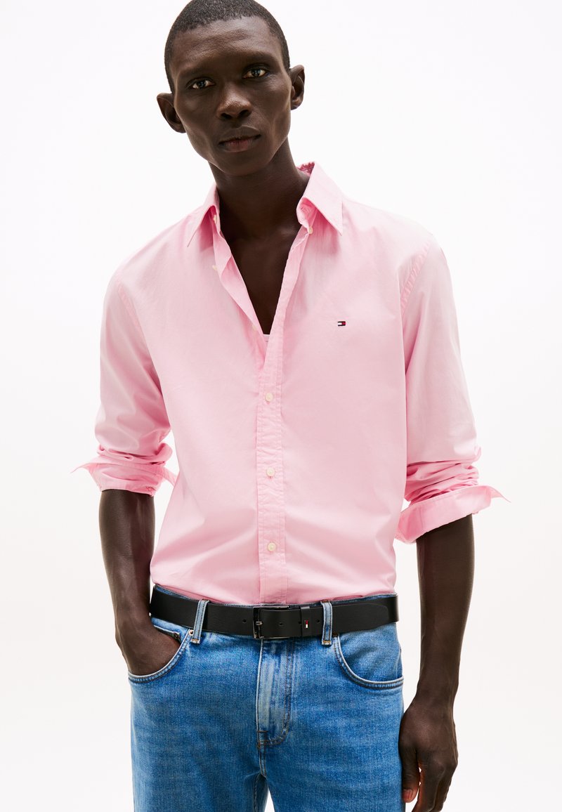 Lys rosa skjorte med knapper og oppbrettede ermer, kombinert med blå denimbukser og svart belte. Viser enkelt design og kontrasterende farger.