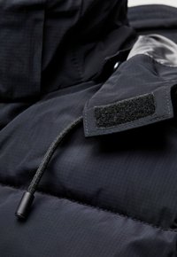 Marineblauer Steppjacke mit strukturiertem Äußeren. Verfügt über einen Kordelzug mit einem schwarzen Stopper und einem Klettverschluss-Patch am Ärmel.