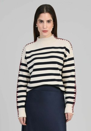 Pull rayé crème avec détails noirs et rouges, col montant côtelé et accents de couture contrastants le long des manches.