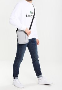 Szara torebka na ramię o gładkiej fakturze, prostokątnym kształcie, regulowanym pasku i wytłoczonym logo. Nosi ją osoba w białej bluzie i jeansach.