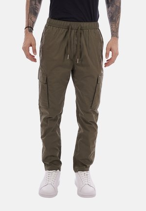 Uomo con pantaloni cargo verde oliva con vita con coulisse e sneaker bianche, in piedi con le braccia rilassate ai lati.