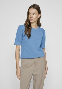 Modrý pletený svetr s krátkým rukávem, s texturovaným vzorem a žebrovanými manžetami, spárovaný s béžovými kalhotami s vysokým pasem. Jednoduchý a uvolněný střih.