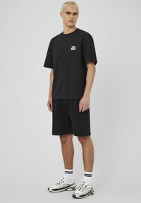 T-shirt en coton noir avec un petit graphique sur la poitrine, associé à un short noir. Porté avec des chaussures de sport argentées et des chaussettes rayées.