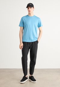 Blaues Baumwoll-T-Shirt, kurze Ärmel, runder Ausschnitt; schwarze tapered Hose; schwarze Sneakers mit weißen Sohlen; schwarze Mütze mit Logo.