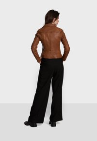 Veste en cuir marron au design ajusté, avec des coutures visibles et un col cranté. Portée sur un pantalon large noir et des chaussures noires.