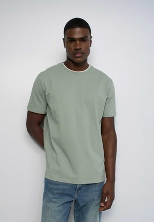 Joven con el pelo corto que lleva una camisa de manga corta de color verde claro y vaqueros azules, de pie frente a un fondo liso y claro.