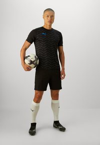 Camiseta deportiva negra con un patrón en forma deChevron, combinada con pantalones cortos negros. Calcetines blancos, botines negros y un balón de fútbol marcado con marca.