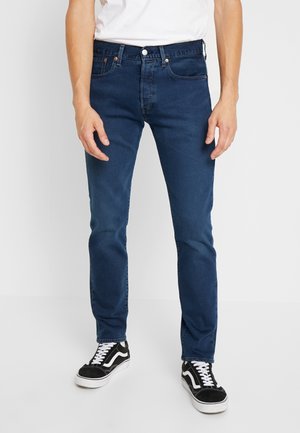Vaqueros oscuros ajustados, hechos de mezclilla, con bolsillos frontales, cierre de botón y detalles de costura en color marrón. Combinados con zapatillas negras.