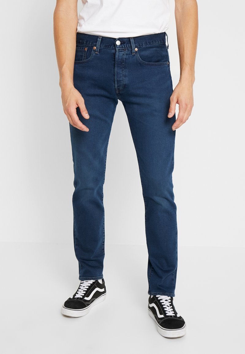 Mörkblå jeans med slim fit, tillverkade av denim, med framfickor, knappgylf och beige sömnadsdetaljer. Bärs med svarta sneakers.