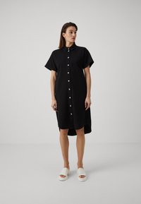 Svart skjorte-kjole med korte ermer, krage, og sidesplit. Hvite slip-on sko med tykk såle. Glatt stoffoverflate.