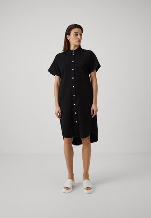 Black button-up shirt dress with short sleeves, collar, and side slit. White slip-on shoes with a chunky sole. Smooth fabric texture.