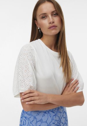 Donna con capelli lunghi che indossa una maglia bianca con maniche a sbuffo e occhielli e una gonna blu a fiori, braccia incrociate, espressione neutra, sfondo chiaro.