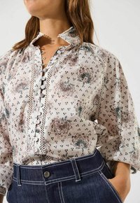 Hvid blomsterbluse med høj krave, blondekant og knaplukning foran. Har indviklede mønstre i grøn og pink. Kombineret med mørk denim.