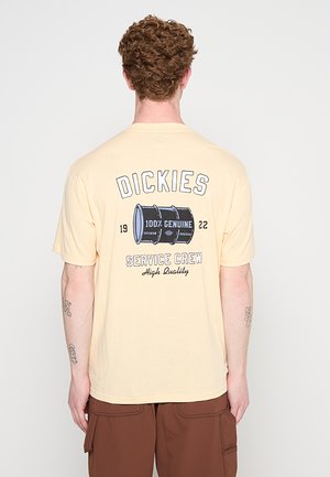 Hombre con cabello rizado que lleva una camiseta beige de Dickies con un gráfico de barril y el texto "100% Genuine Service Crew High Quality" en la espalda.