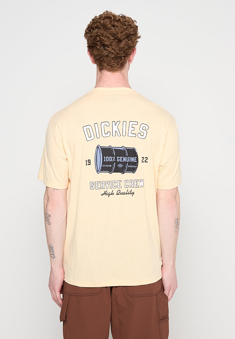 Uomo con capelli ricci che indossa una maglietta beige Dickies con una grafica di una botte e il testo "100% Genuine Service Crew High Quality" sul retro.