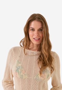 Maglione leggero color crema a maglia con trama aperta, caratterizzato da motivi colorati sul petto. Maniche lunghe e scollo tondo.