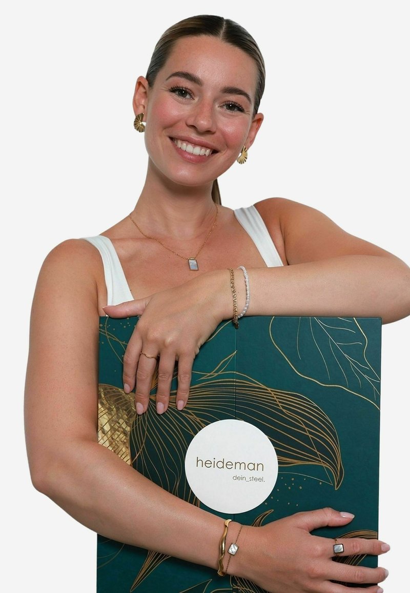 A woman holds a green and gold box with "heideman dein_steel." The box features leaf patterns and a glossy texture. Jewelry includes a gold bracelet.