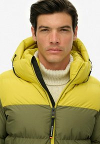 Joven con cabello oscuro que lleva una chaqueta de plumas verde y amarilla sobre un suéter blanco de cuello alto, mirando hacia adelante con una expresión neutral.