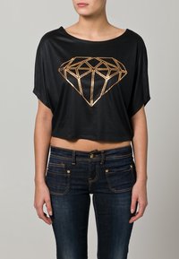 Černé zkrácené tričko s designem zlatého geometrického diamantu. Kombinováno s tmavě modrými džíny s předními kapsami a zlatými akcenty.