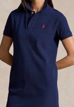 Kvinde iført en navyblå polo-kjole med korte ærmer og en rød broderet logo på brystet, stående med hænderne bag ryggen.