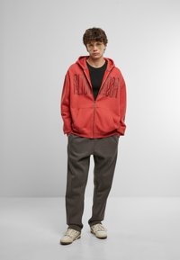 Red zip-up hoodie with front pockets and embossed logo, paired with loose gray sweatpants and light-colored sneakers. Model wears glasses.