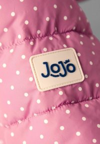 Chaqueta de plumas rosa con lunares blancos, que presenta una etiqueta texturizada beige con letras azules que dice "JoJo" de manera prominente.