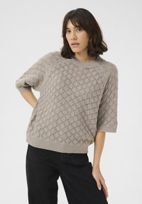 Jasno beżowy sweter dzianinowy z teksturowanym wzorem w diamenty, kr�ótkimi rękawami i elastycznym ściągaczem na dole. W połączeniu z czarnymi dżinsami.