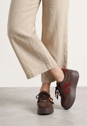 Person, die beige verkürzte Hosen und braune Plateausneaker mit roten Streifen und dekorativen Perlen auf den Schnürsenkeln trägt, steht auf einem hellen Boden.